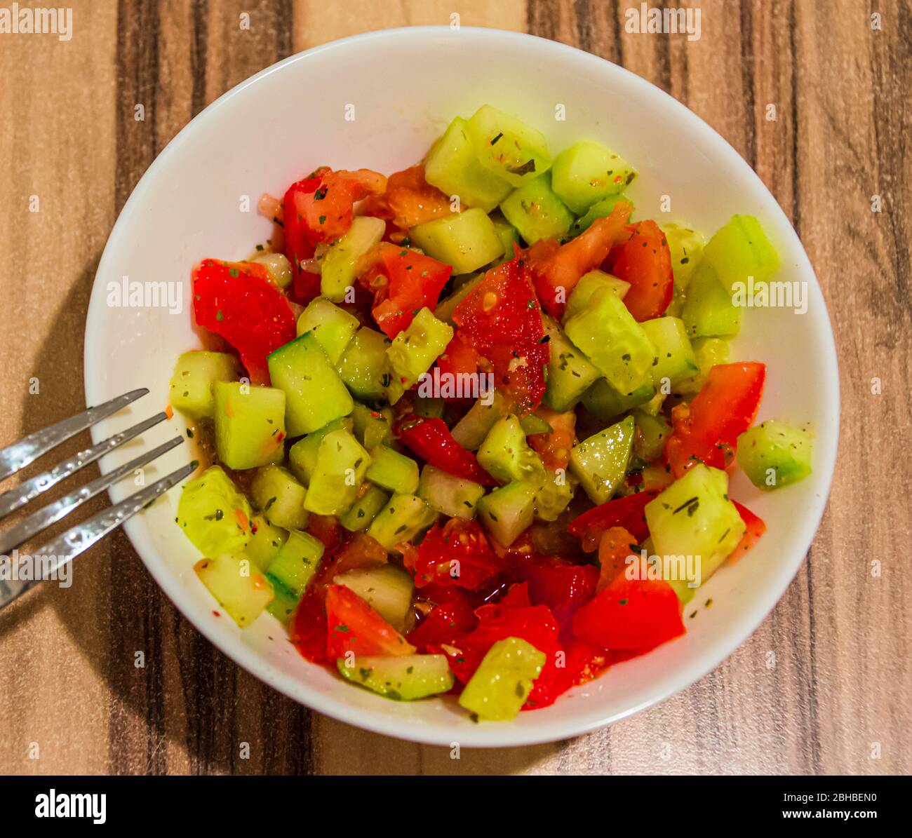 Vista dall'alto dell'insalata succosa di pomodori e cetrioli preparati con spezie in una ciotola bianca con una forchetta sul lato sinistro Foto Stock