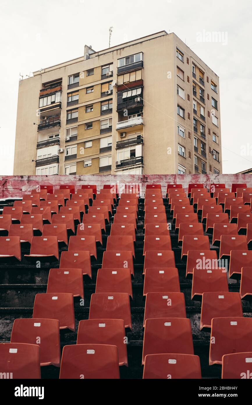 File di posti a sedere rossi vuoti nello stadio con architettura Brutalista, alto edificio di edifici di appartamenti sullo sfondo Foto Stock