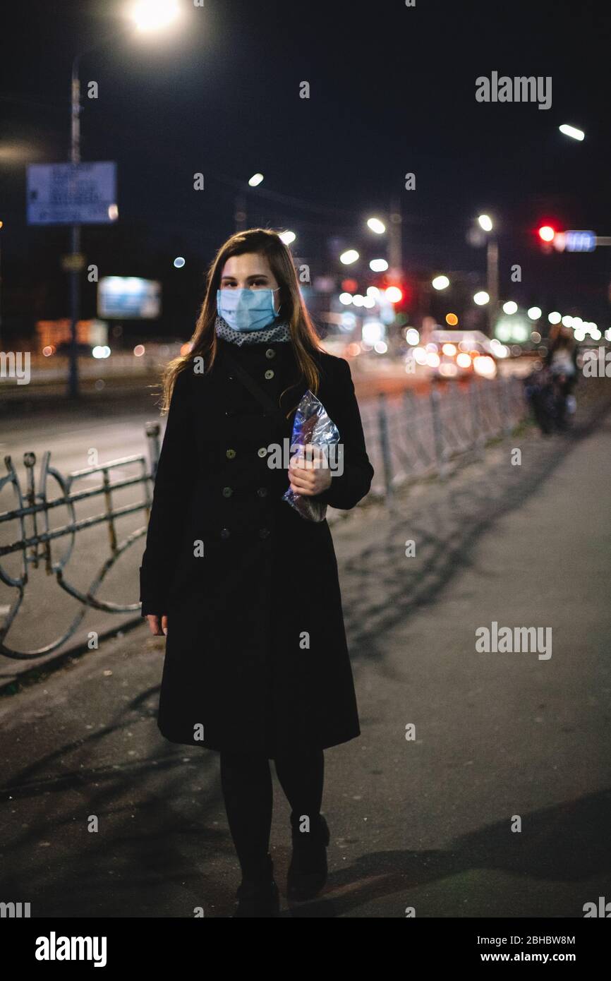 Giovane donna che indossa maschera medica con il viso e il pane mentre cammina per la strada della città di notte Foto Stock