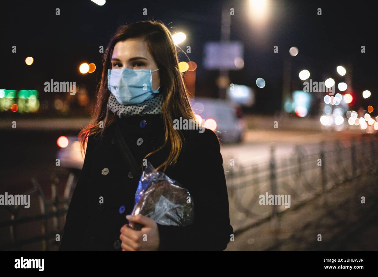 Ritratto di giovane donna che indossa maschera medica viso che porta il pane mentre camminano sulla strada della città di notte Foto Stock