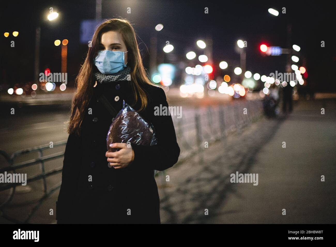 Ritratto di donna giovane triste che indossa faccia maschera medica che porta il pane mentre cammina sulla strada della città di notte Foto Stock