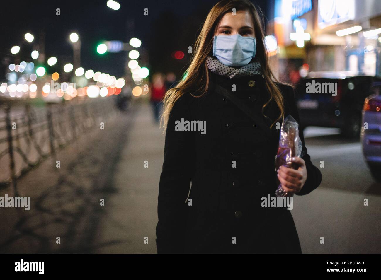 Giovane donna che indossa maschera medica con il viso e il pane mentre cammina per la strada della città di notte Foto Stock