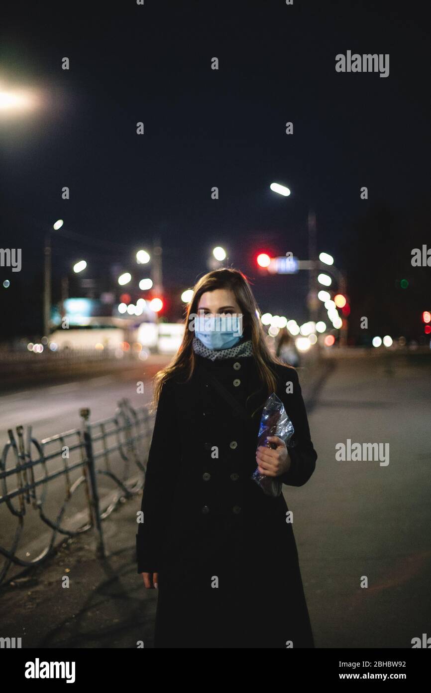 Giovane donna che indossa maschera medica con il viso e il pane mentre cammina per la strada della città di notte Foto Stock