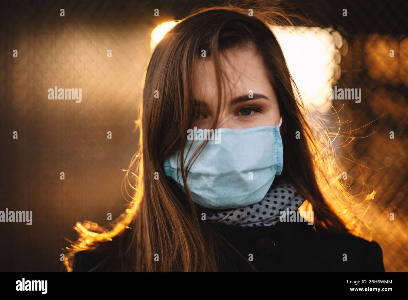 Ritratto di giovane donna che indossa maschera protettiva viso medico mentre in piedi contro recinto al tramonto Foto Stock
