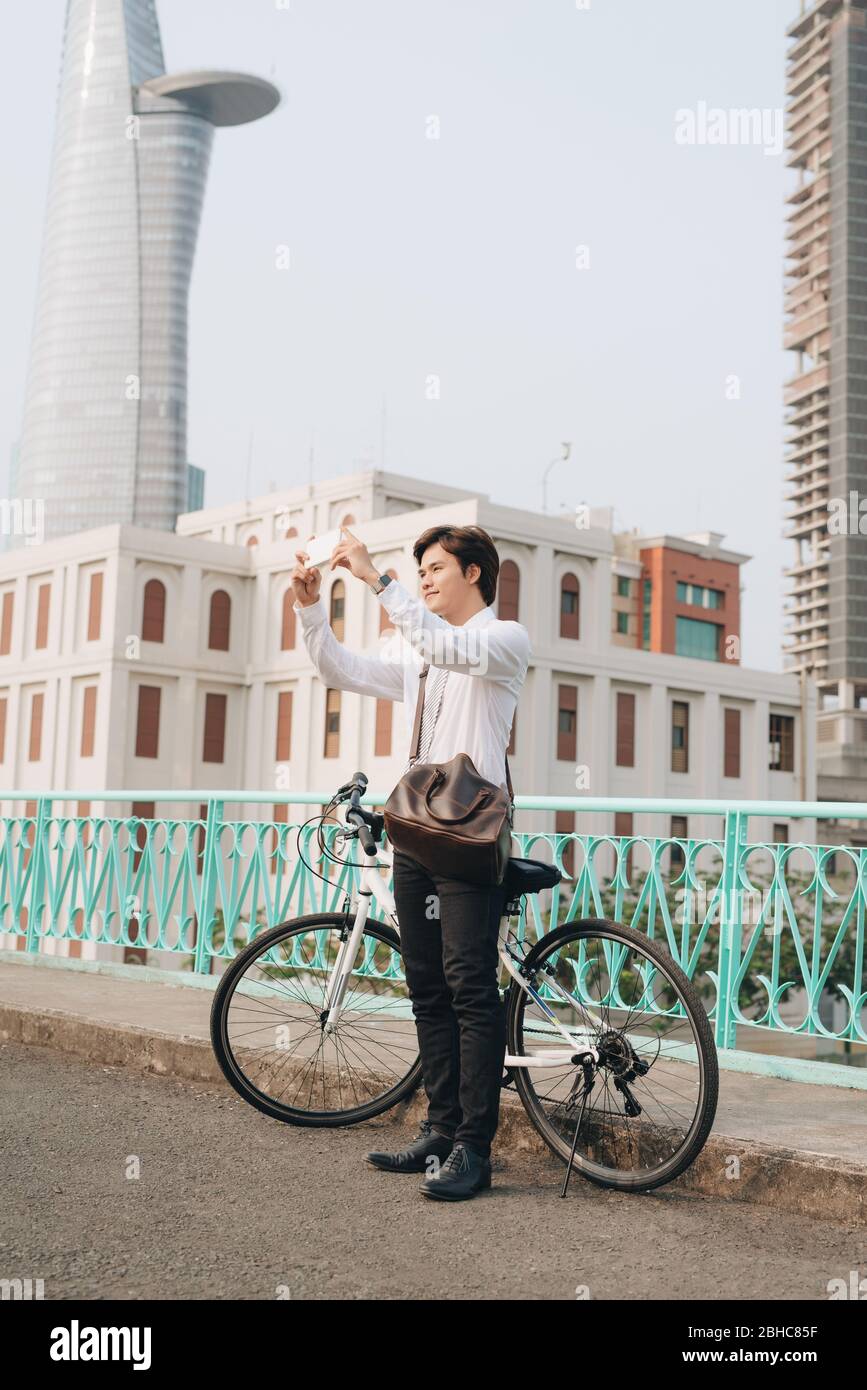 Giovane uomo di scattare una foto con il suo smart phone mentre in piedi sulla strada di città con la sua bicicletta accanto a lui Foto Stock