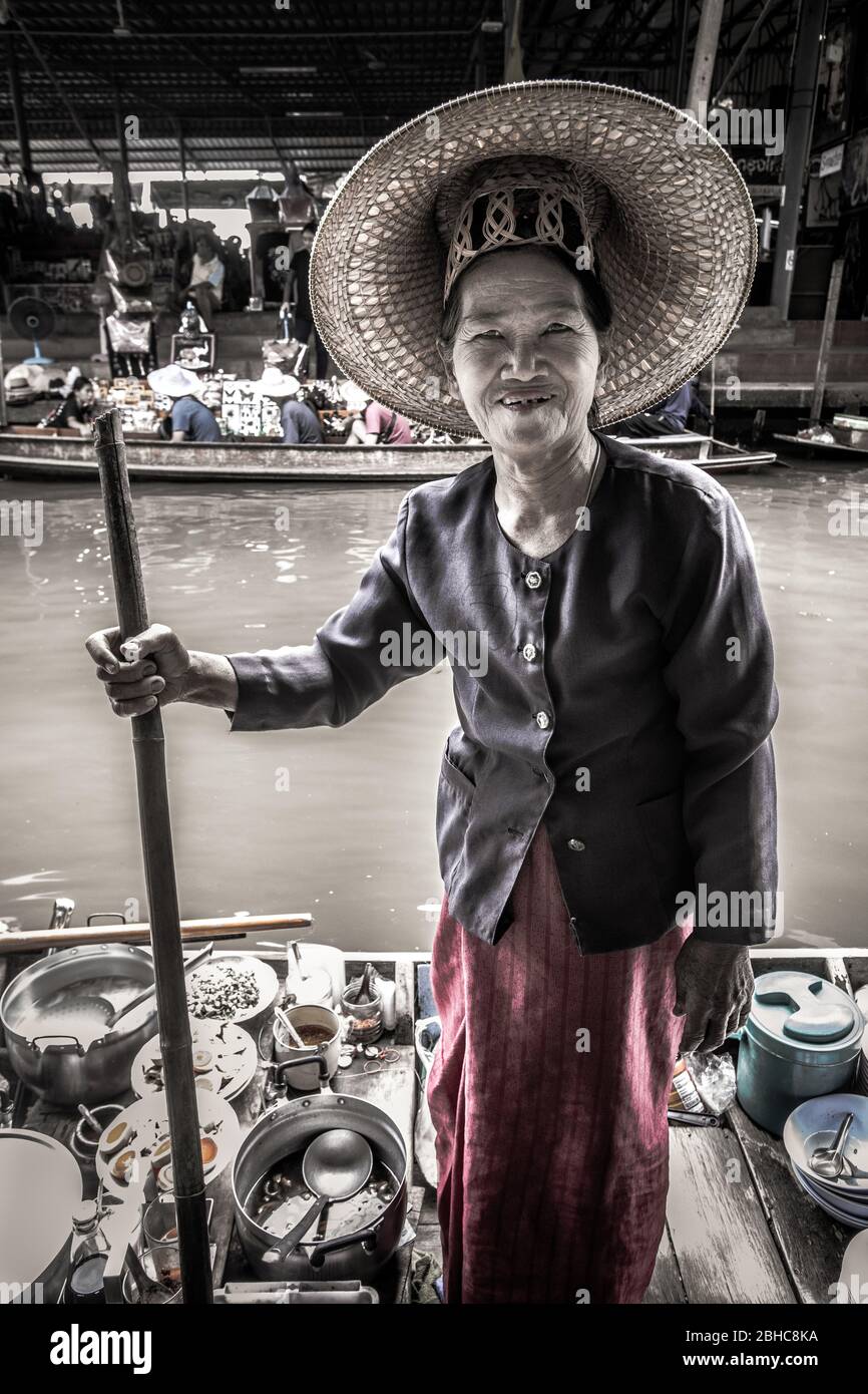 Damnoem Saduak, Thailandia. Mercato galleggiante. Anziana signora tailandese con tipico cappello locale su una piccola barca che vende i suoi prodotti vegetali. Cercando e smilin Foto Stock