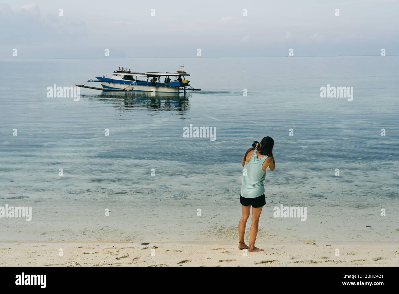 Ragazza fotografando una barca locale sulla spiaggia quando il colore del mare si mescola con il cielo in un paradiso terreno. Colori pastello e delicati. Indonesia i Foto Stock