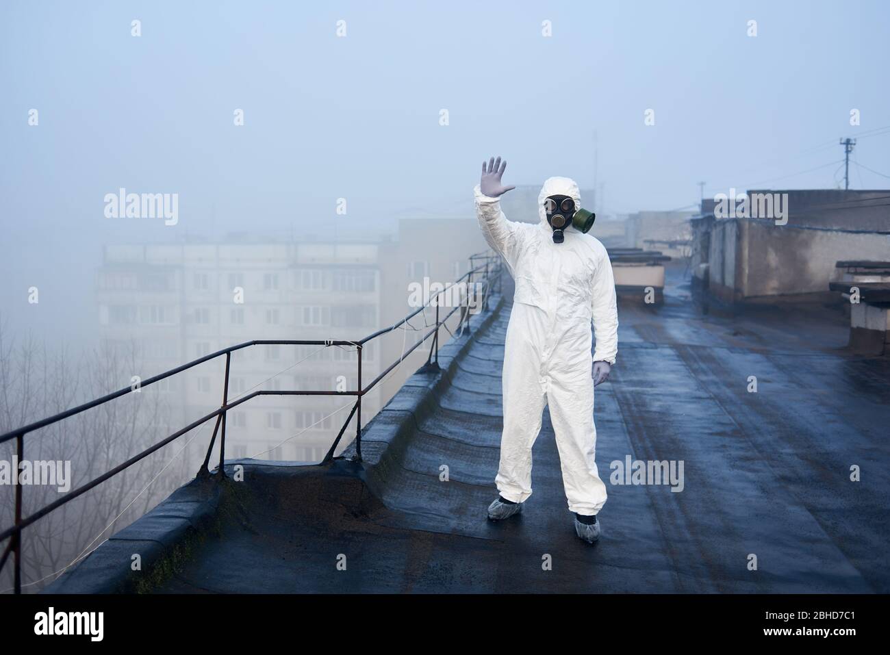 Scienziato in piedi sul tetto di blocco di appartamenti, mostrando stop, indossando tuta di protezione bianca e maschera, esplorando l'ambiente per la ricerca futura Foto Stock