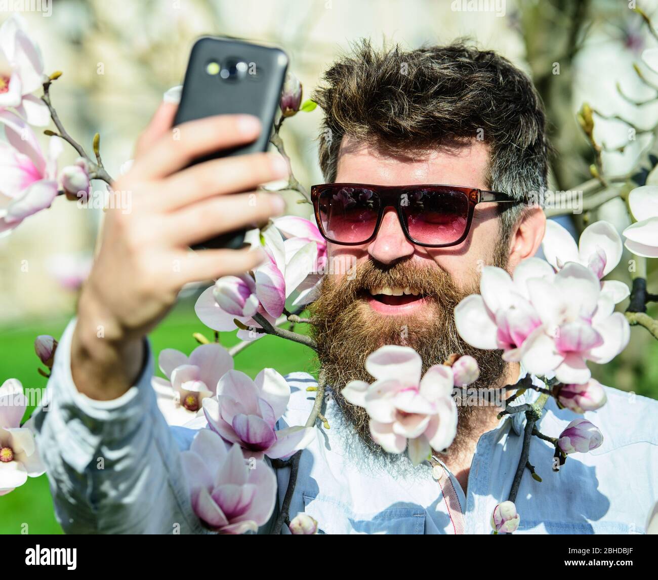 Hipster felice in occhiali da sole eleganti, scattare foto selfie,  streaming video su smartphone. L'uomo con barba e baffi indossa occhiali da  sole il giorno di sole, fiori magnolia sullo sfondo. Concetto