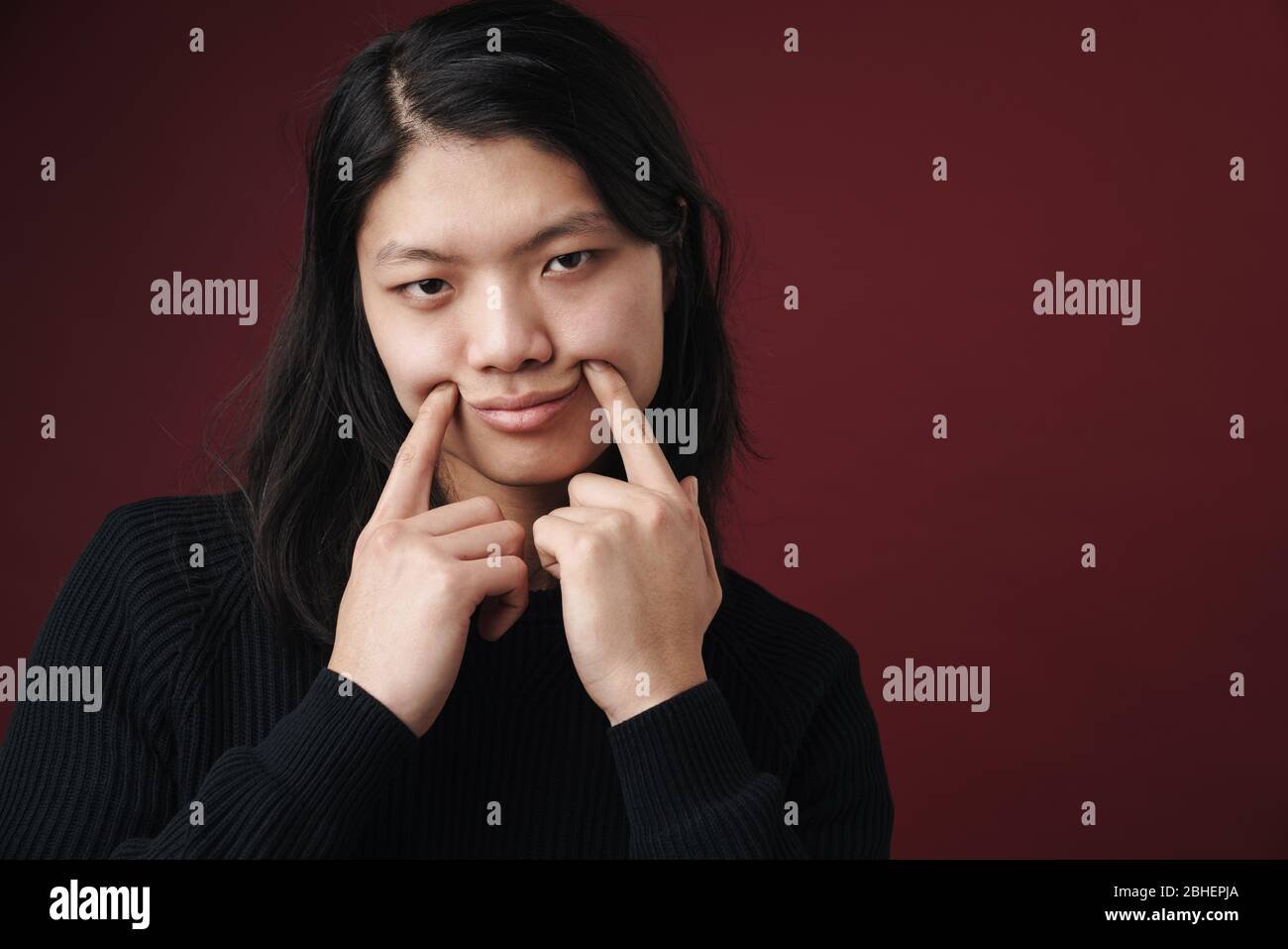Immagine di un uomo asiatico triste che fa un sorriso falso con le dita isolate sullo sfondo borgogna Foto Stock