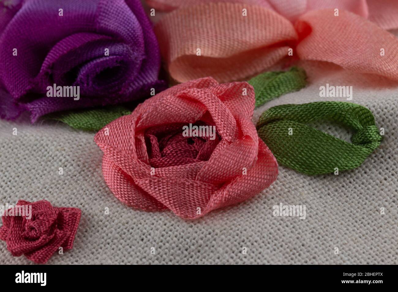 macro ripresa di una varietà di rose da ricamo a nastro di seta su tela. Nelle tonalità rosa, rossa, malva e viola con foglie verdi Foto Stock