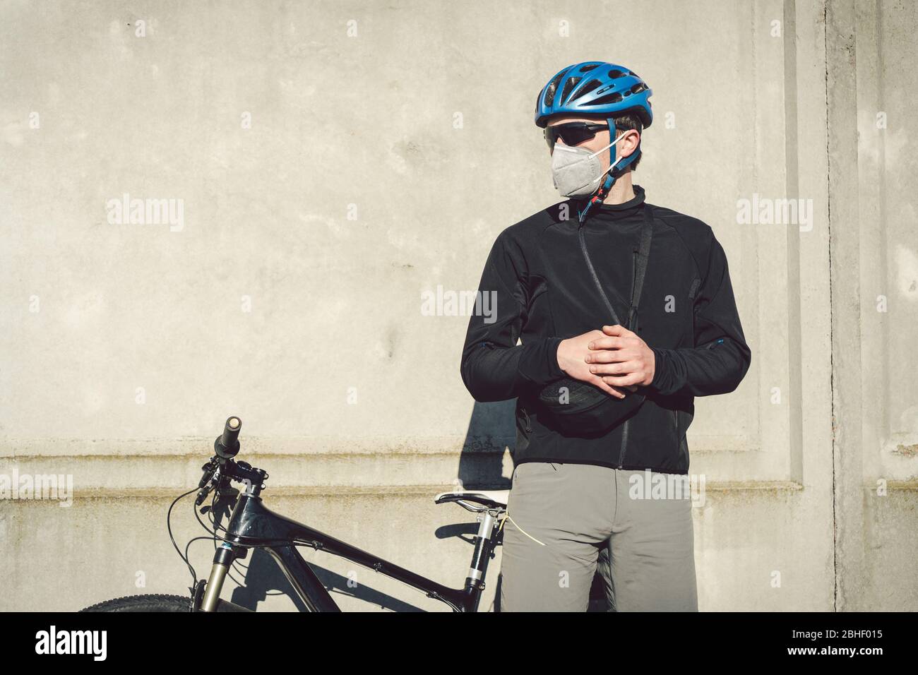 Un ciclista uomo in una maschera facciale protettiva si trova accanto ad una bicicletta su uno sfondo di parete grigia di cemento. Corriere non riconoscibile nel respiratore. Consegna Foto Stock