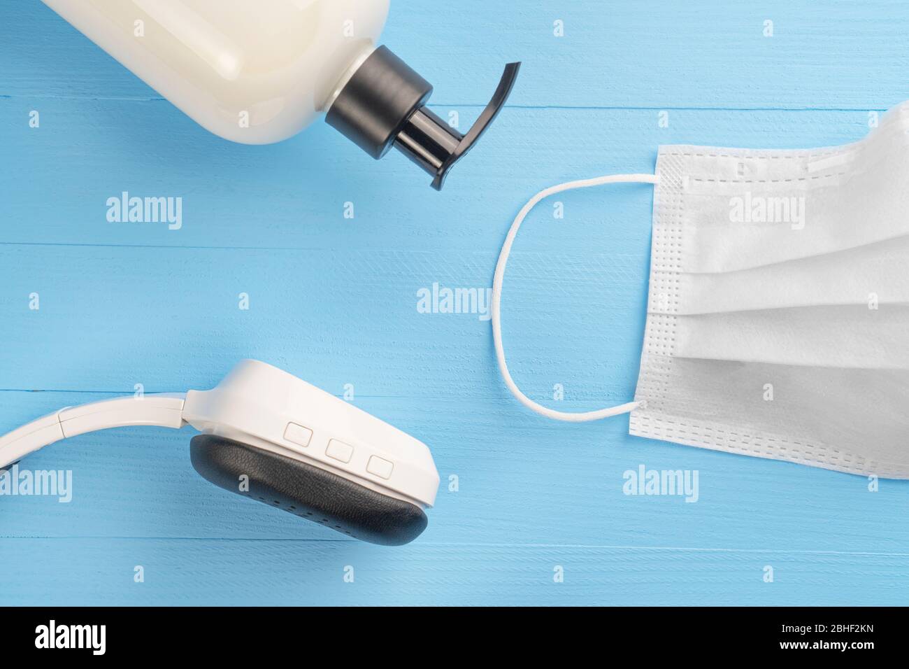 Maschera bianca per uso medico con contenitore per sapone in gel beige e cuffie wireless bianche nere su sfondo blu chiaro in legno. Il concetto di soggiorno a casa Foto Stock