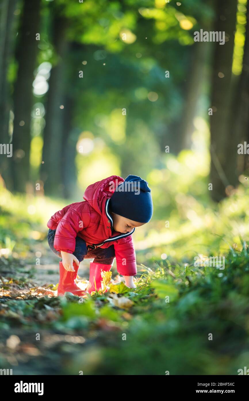 Bambino piccolo in stivali di gomma rossi e giacca rossa nel parco primaverile. Lussureggiante foresta foglie sullo sfondo Foto Stock