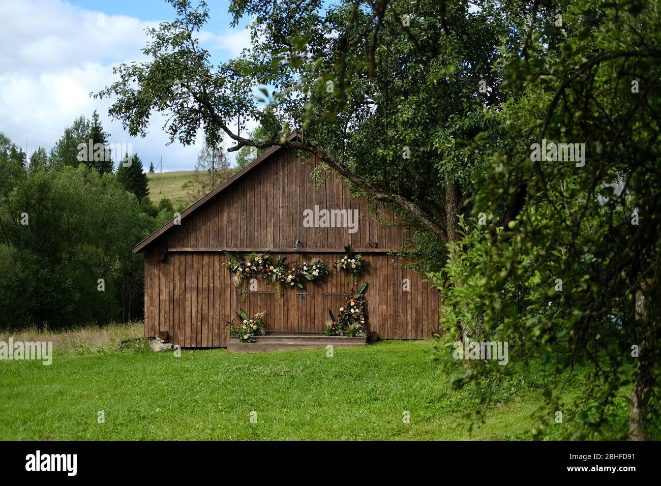 Vecchio fienile marrone parete sfondo legno circondato da alberi verdi. Il fienile è decorato con fiori e verde. Foto luminosa della campagna. Foto Stock