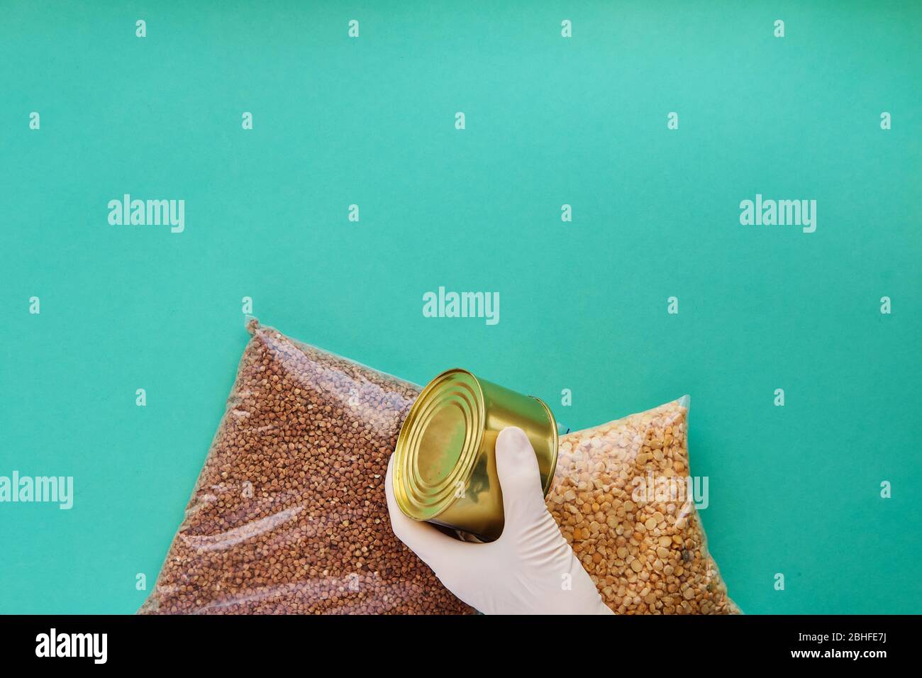 Una mano in un guanto medico tiene il cibo in scatola, accanto ad esso sono pacchetti di cereali su uno sfondo blu Foto Stock