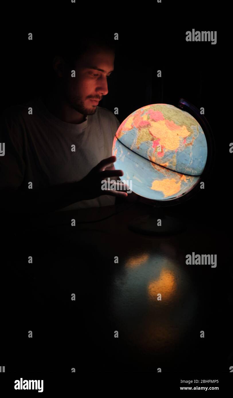 Un uomo che guarda un globo rotante Terra lampada luce illuminata scrivania Mappa Geografia nella notte buia con il volto illuminato Foto Stock