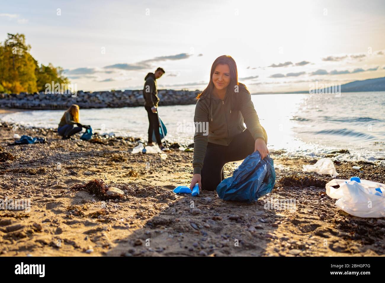 Sorridente giovane donna che pulisce la spiaggia con volontari durante il tramonto Foto Stock