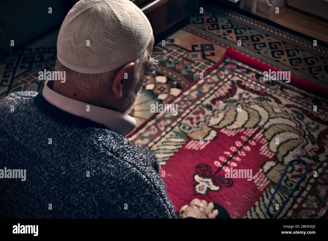 Molto vecchio maschio turco-musulmano barbuto ai suoi anni 80 indossando un cappuccio di preghiera facendo preghiera Salah a casa sua sul tappeto di preghiera nel mese di Ramadan Foto Stock