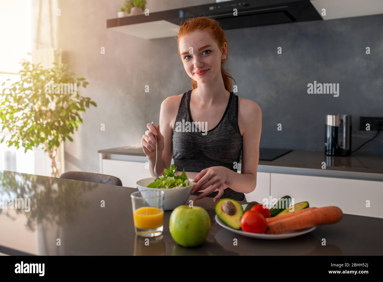 La ragazza atletica con i vestiti della palestra mangia un'insalata nella cucina Foto Stock