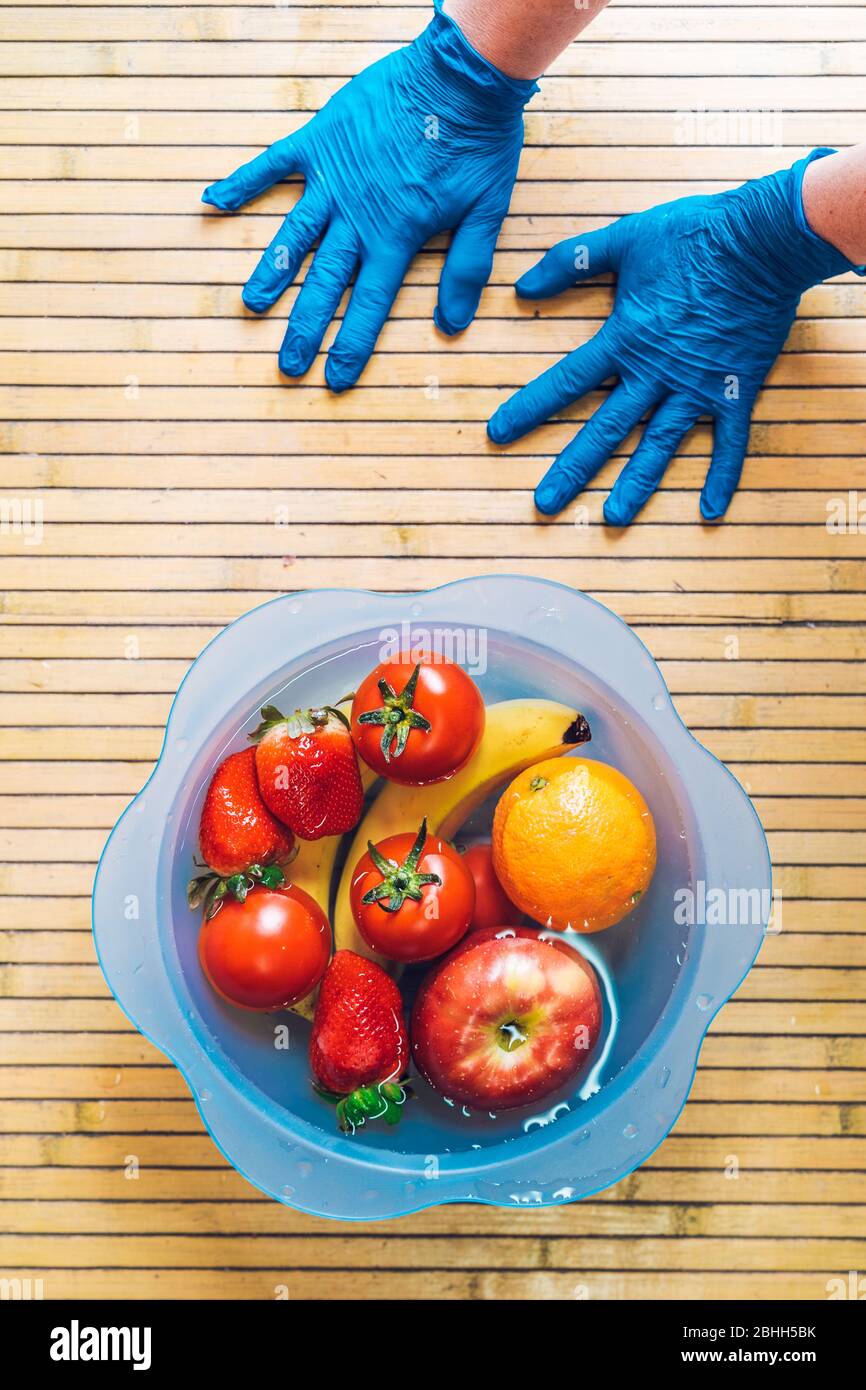 Mani con guanti in lattice blu e una ciotola blu con frutta fresca e pulita su una base di legno. Banane, pomodori, mele, fragole e orang Foto Stock
