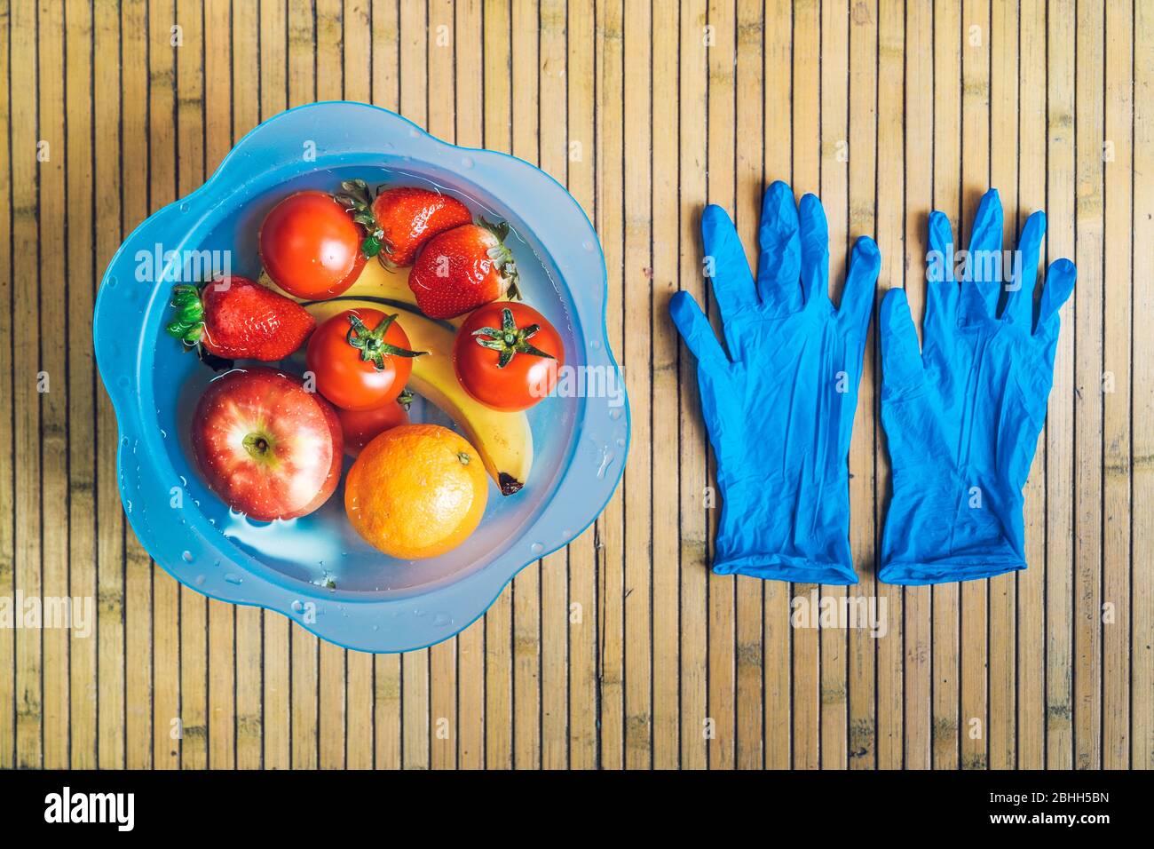 Ciotola blu con frutta fresca e pulita su una base di legno con guanti in lattice blu. Banane, pomodori, mele, fragole e arance immerse Foto Stock