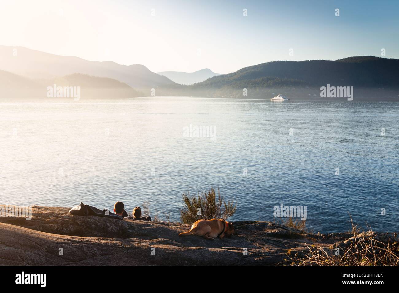 Una coppia felice nell'amore che si rilassa con il loro cane guardando il tramonto con una vista sull'oceano e un'isola. Foto Stock