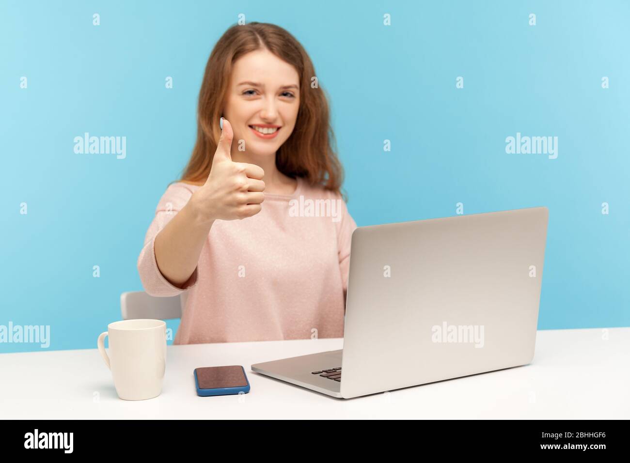 Ottimo lavoro! Donna soddisfatta positiva con un sorriso accattivante e toothy che mostra i pollici su gesto, come, approvazione, lavorando su notebook da casa ufficio. Foto Stock