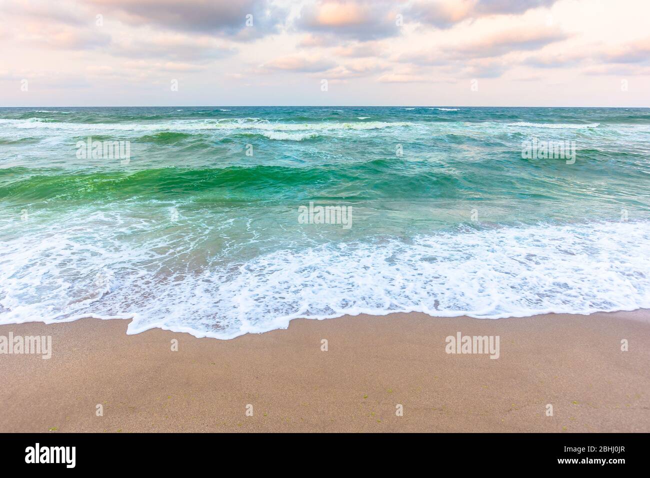 tramonto dorato e nuvoloso sopra le acque verdi del mare. onde che si infrangono sulla spiaggia sabbiosa. orizzonte limpido. tempo ventoso in continuo mutamento Foto Stock