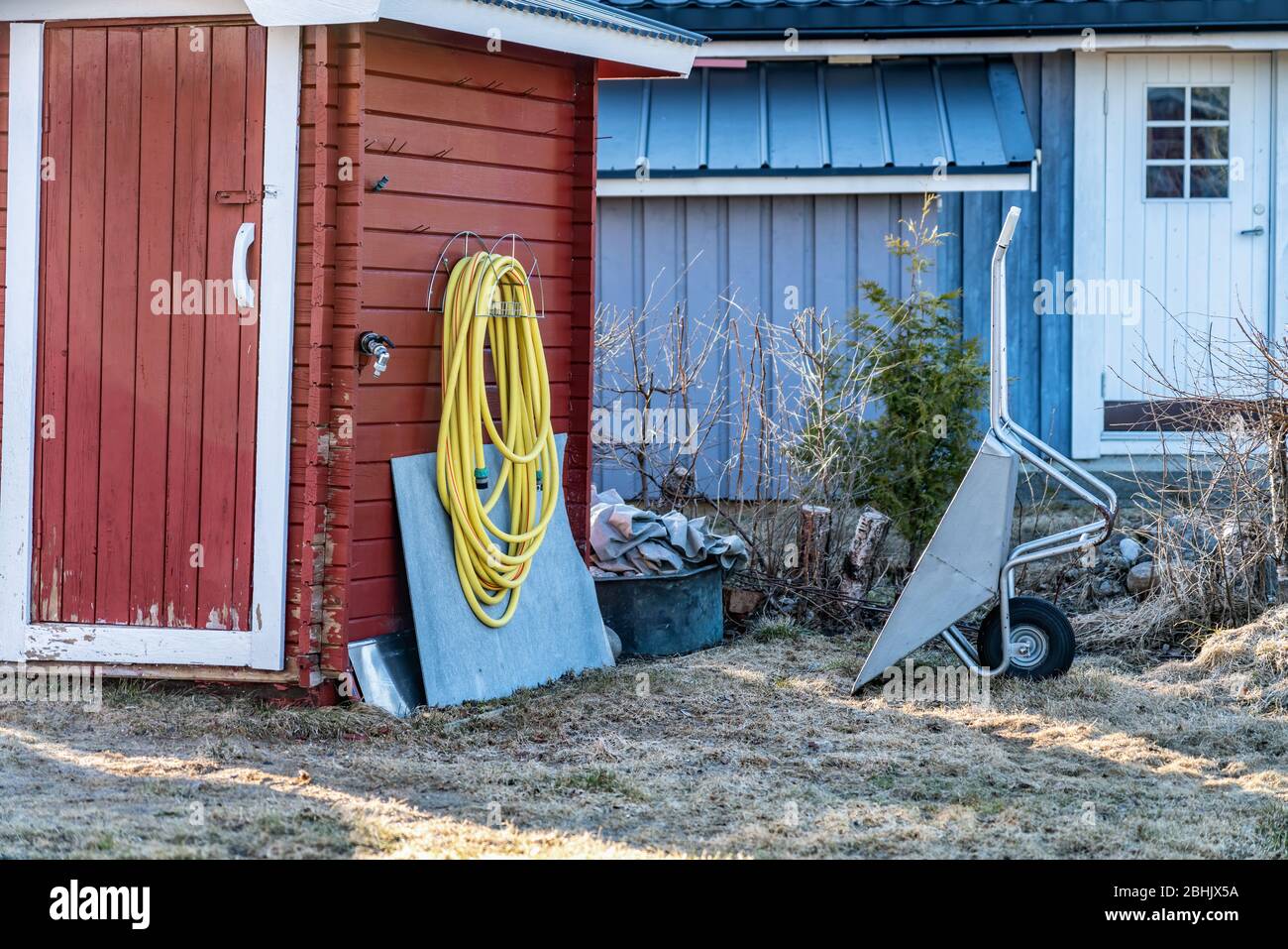 Tubo di irrigazione in gomma gialla per piante appendiabiti su pareti di legno rosso di capanna tradizionale svedese giardino, rubinetto acqua, carriola grigia Foto Stock