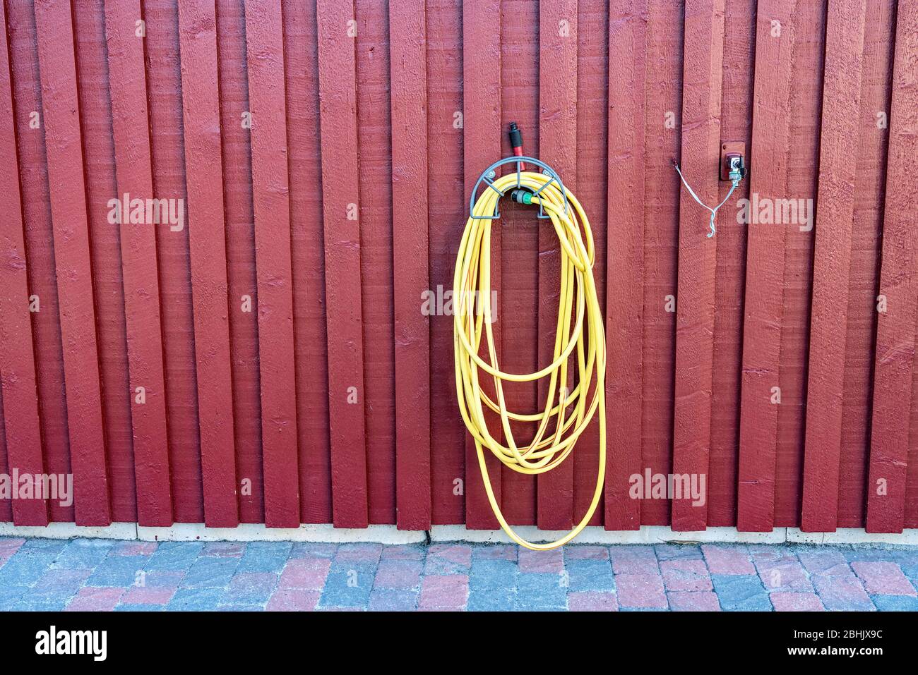Tubo di irrigazione in gomma gialla per piante appesa su parete di legno rosso di tradizionale capannone da giardino svedese vicino al rubinetto d'acqua montato a parete. Foto Stock