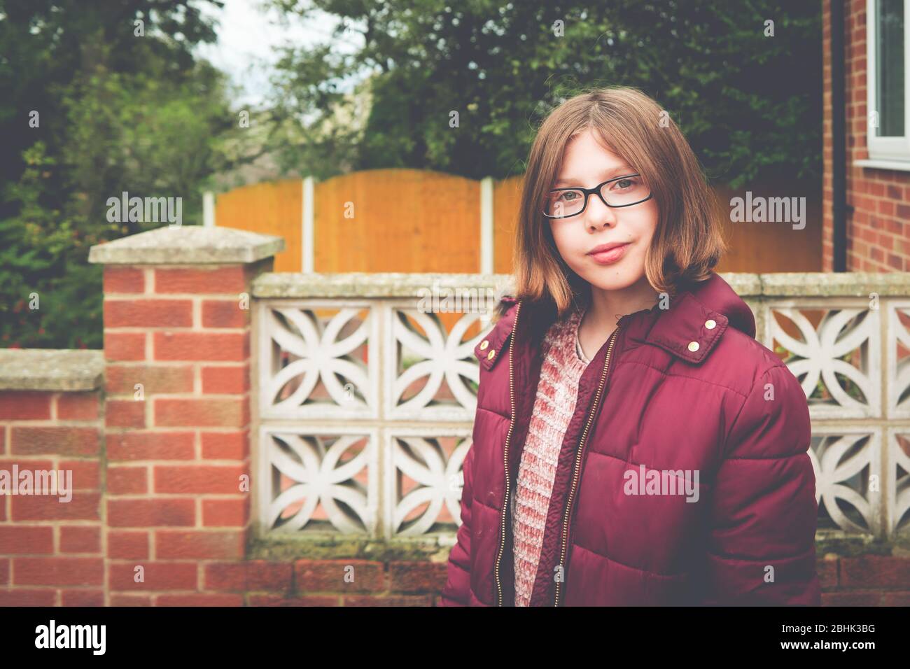 Autentico ritratto suburbano giardino di una ragazza fiduciosa con busto corto, occhiali e spesso cappotto Foto Stock