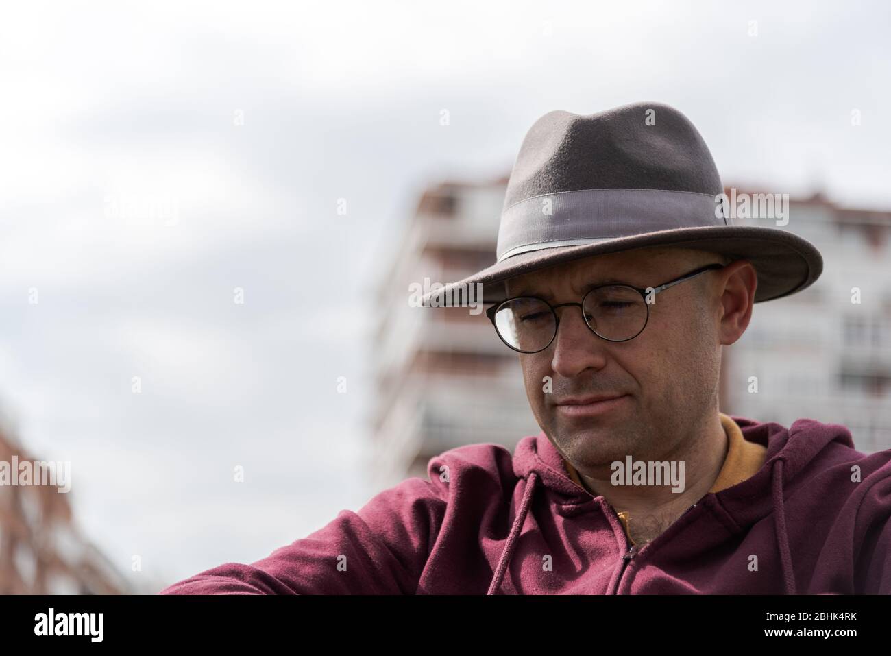 Uomo caucasico con occhiali e cappello depressivo e triste in una sedia a rotelle solitaria in un parco con il cielo e alcuni edifici sullo sfondo con lo spazio Foto Stock