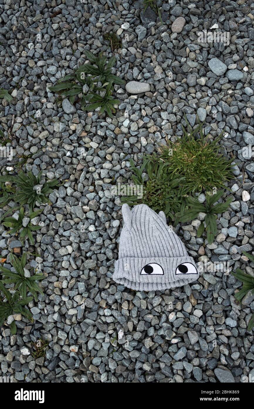 Cappello di lana di lana grigio con occhi, abbandonato e sdraiato a terra circondato da ghiaia e erbacce, visto dall'alto Foto Stock