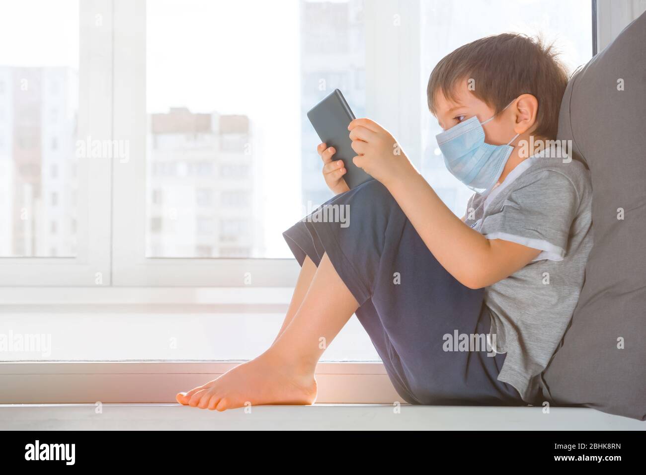 Ragazzo che studia online a casa con un tablet. Covido19 pandemico globale, Coronavirus. Bambino in maschera medica protettiva siede su windowsill e gioca Foto Stock