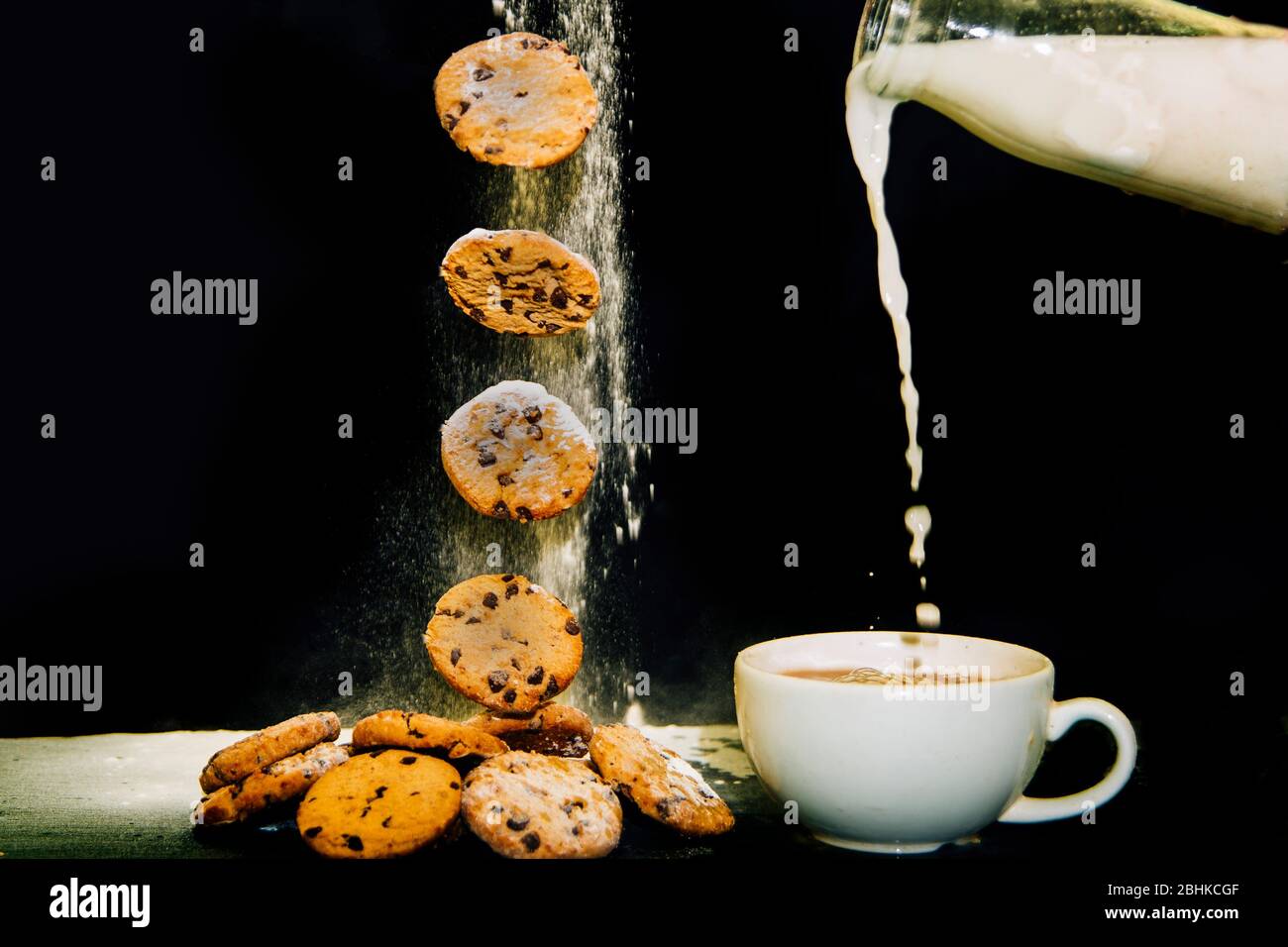 Tè e biscotti. Biscotti o biscotti cadenti su una pila di biscotti accanto ad una tazza di tè su uno sfondo nero. Latte versato in tazza di tè. Foto Stock
