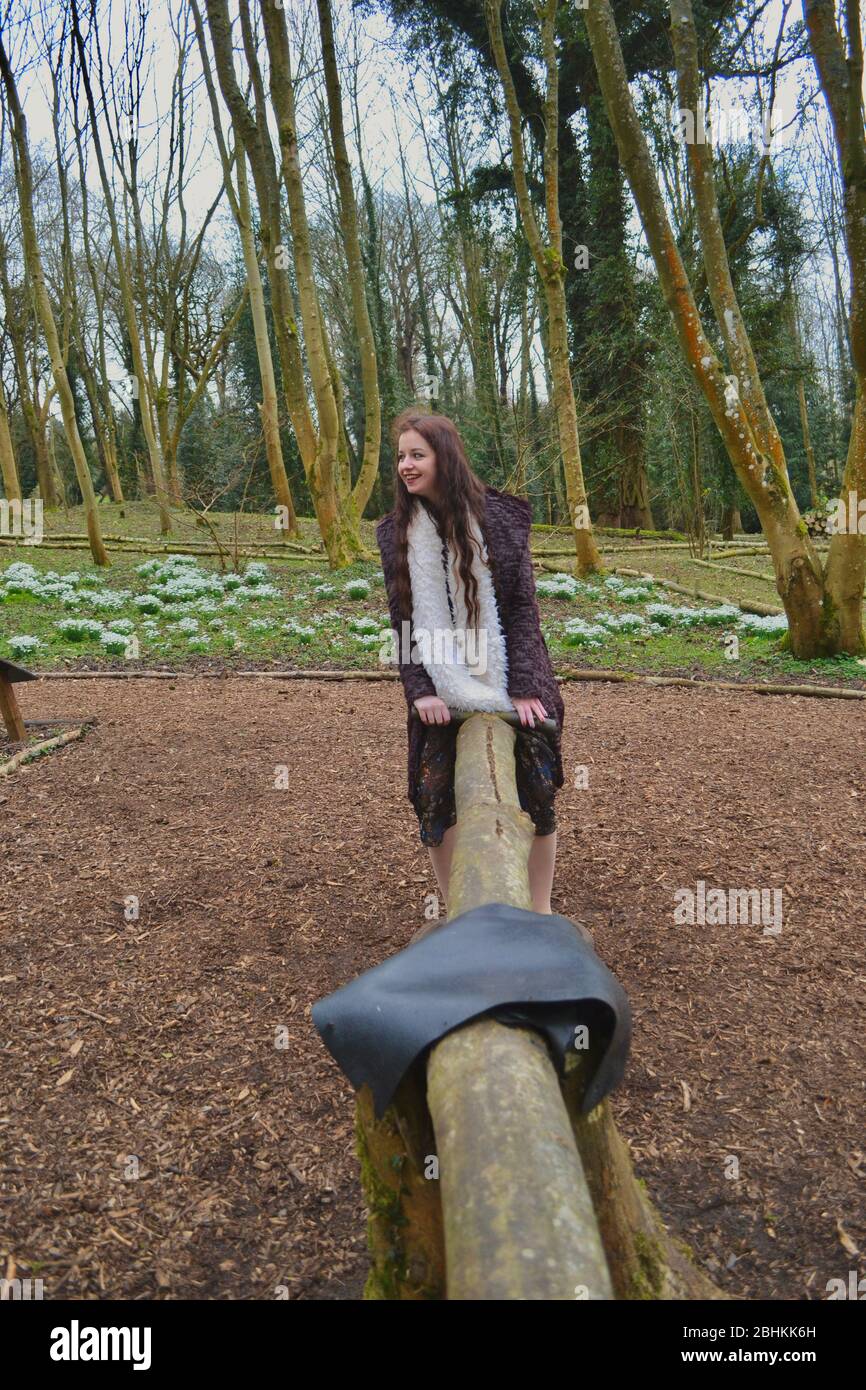 Giovane ragazza felice con lunghi capelli brunette e un cappotto e sciarpa, giocando su una vista rustica in un parco giochi nel bosco in inverno Foto Stock