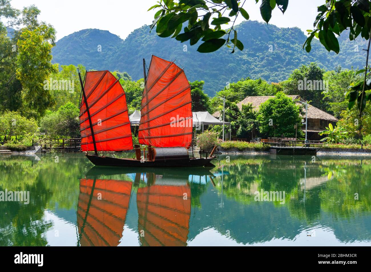 Spazzatura. Bella barca tradizionale vietnamita con vele rosse su un pittoresco lago nella giungla. Vele rosse riflesse nell'acqua. Foto Stock