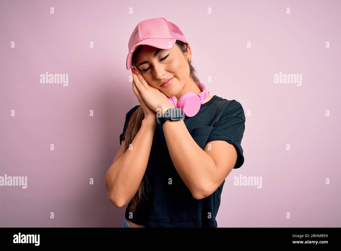 Giovane bella sport fitness donna che indossa cappuccio da allenamento e cuffie su sfondo rosa dormire stanco sognare e posare con le mani insieme w Foto Stock