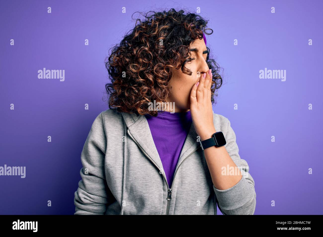 Bella ragazza sportiva curly arab fare sport indossare abbigliamento sportivo su sfondo viola annoiato yawning stanco copertura bocca con mano. Restless e slee Foto Stock