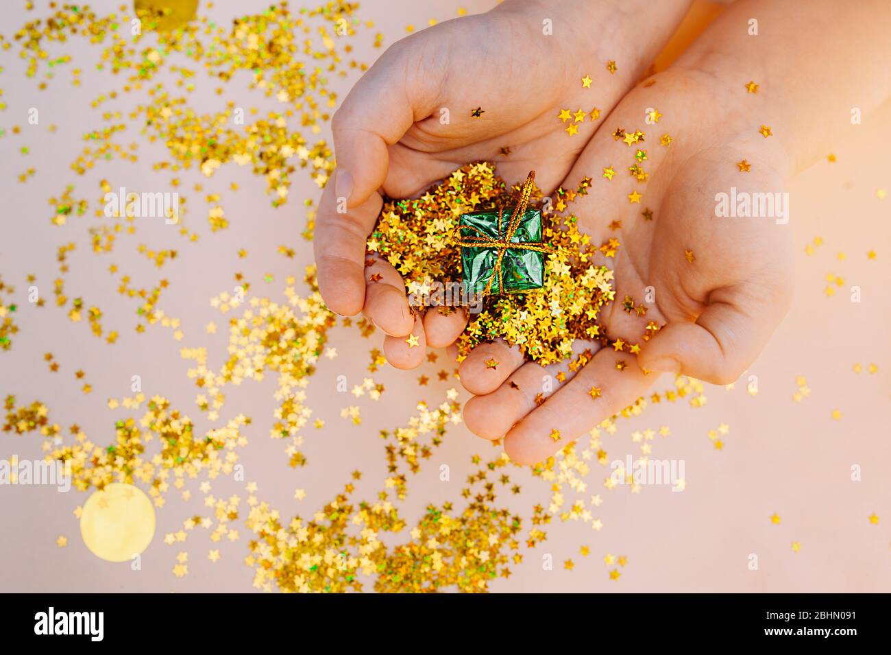 le mani con pelle chiara tengono un piccolo regalo in lamina verde su uno sfondo di stelle d'oro sparse su uno sfondo beige delicato. Foto Stock