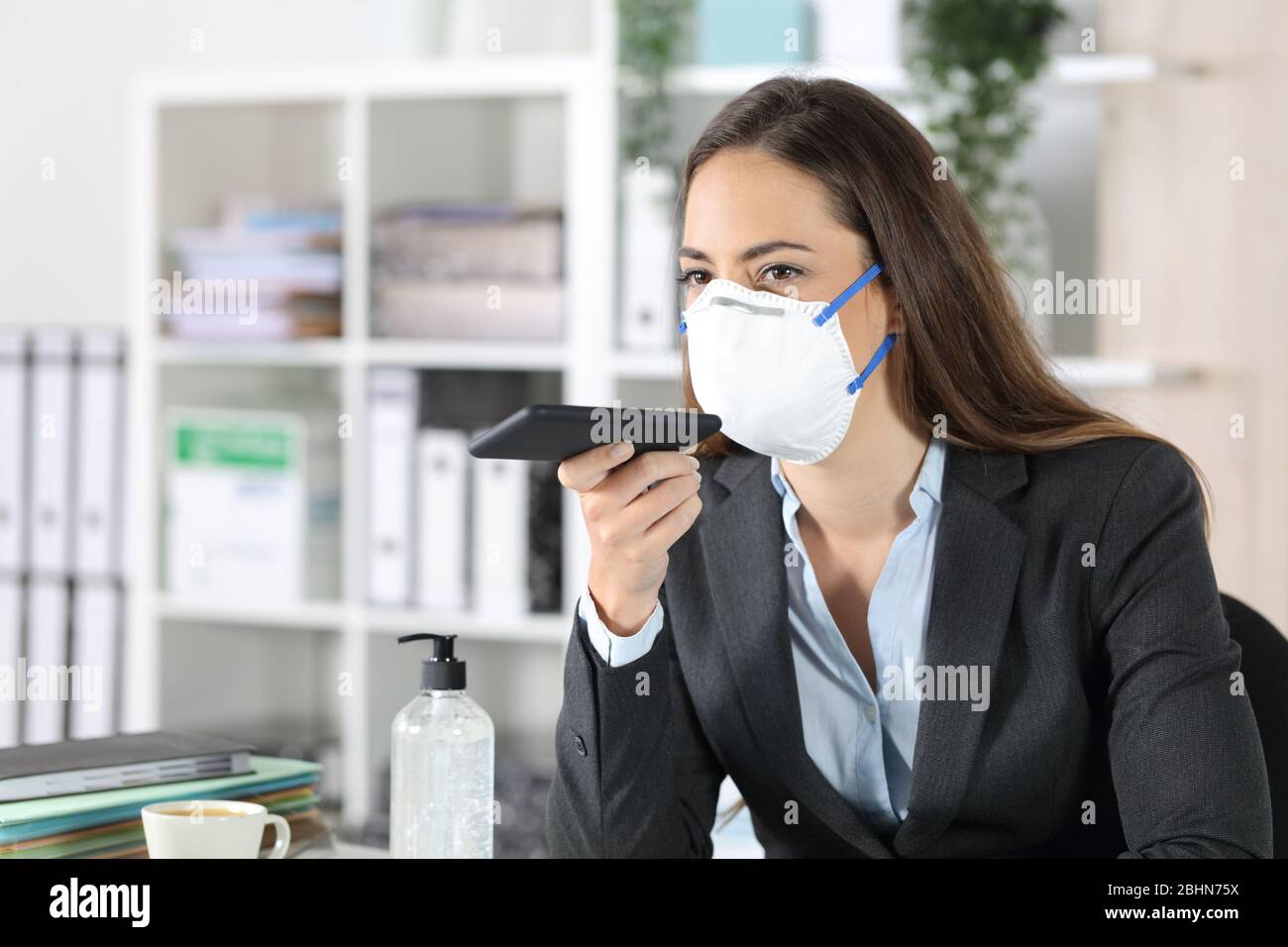 Donna executive con maschera protettiva che utilizza il sistema di riconoscimento vocale sullo smartphone in ufficio Foto Stock