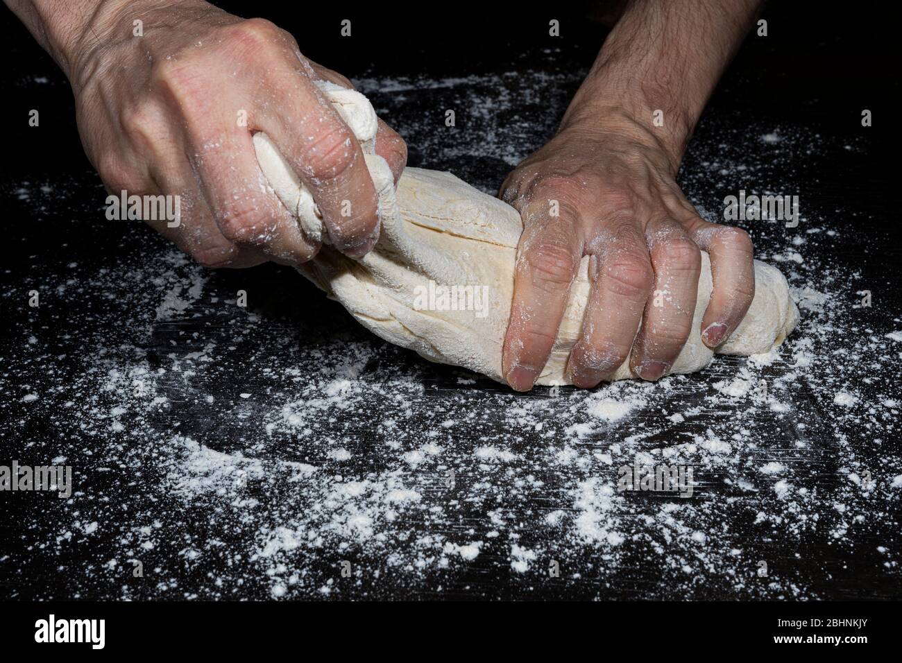 Le mani di un uomo separano un pezzo di pasta da una palla impastata con farina per fare il pane su una superficie nera piena di farina Foto Stock
