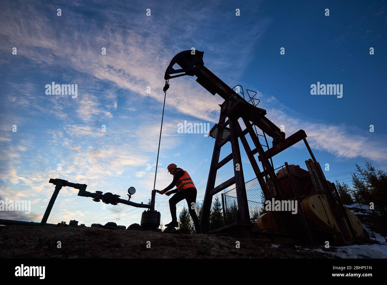 Vista laterale silhouette di Oil MAN in giubbotto da lavoro e casco che  ripara il martinetto della pompa di petrolio. Lavoratore petrolifero che  utilizza la pompa a bilanciere in campo petrolifero sotto