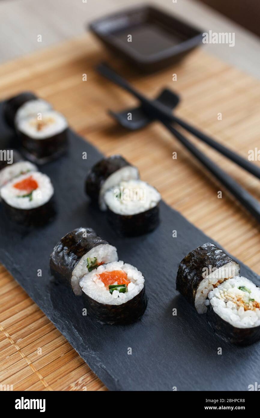 Sushi maki su una lavagna nera. Salsa di soia in un piatto e bastoncini neri su un tappetino di bambù. Foto Stock