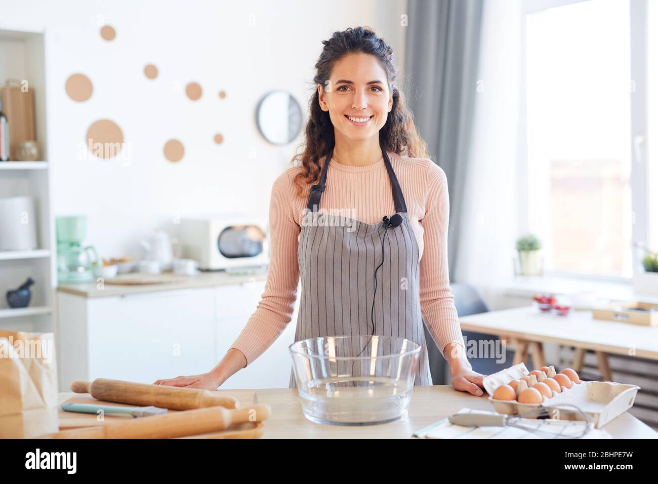 Orizzontale ritratto medio di giovane donna allegra che indossa grembiule in piedi al tavolo da cucina guardando la macchina fotografica sorridente Foto Stock