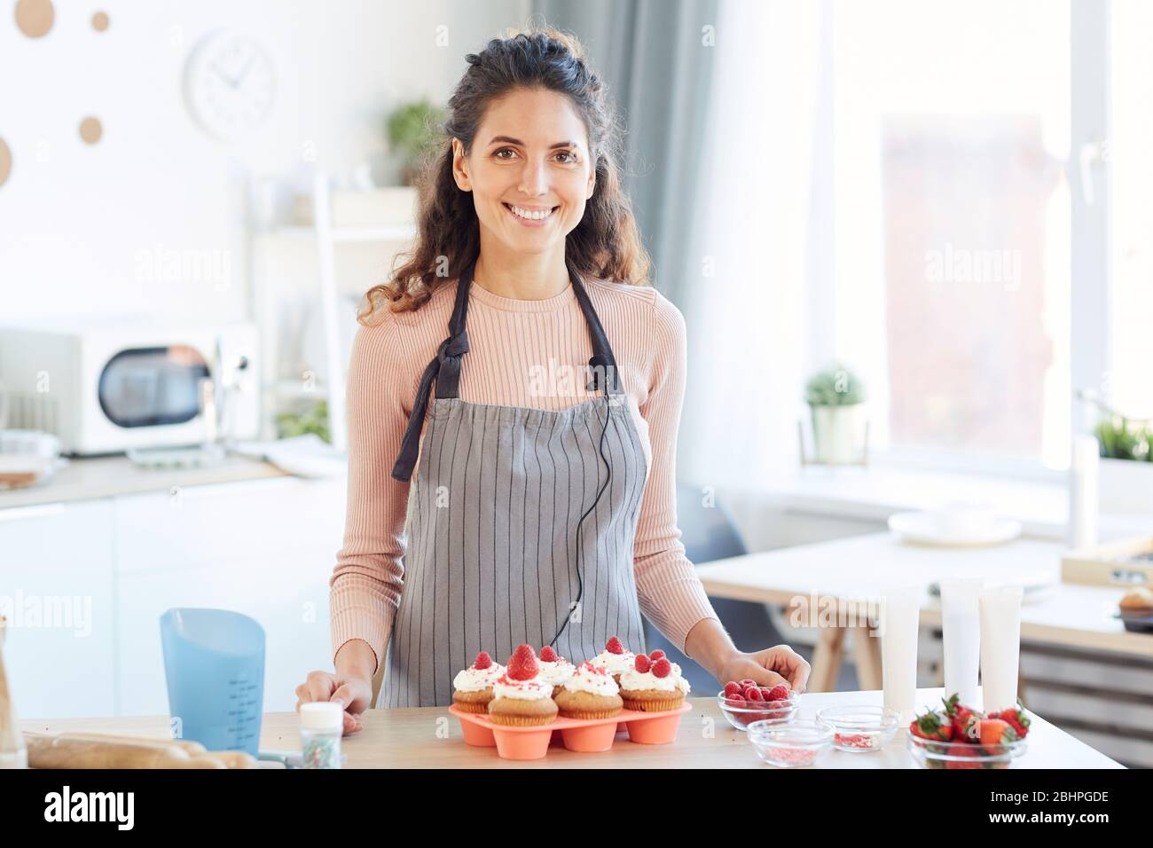 Ritratto orizzontale medio di gioiosa donna caucasica in piedi al tavolo da cucina con cupcake fatti in casa Foto Stock