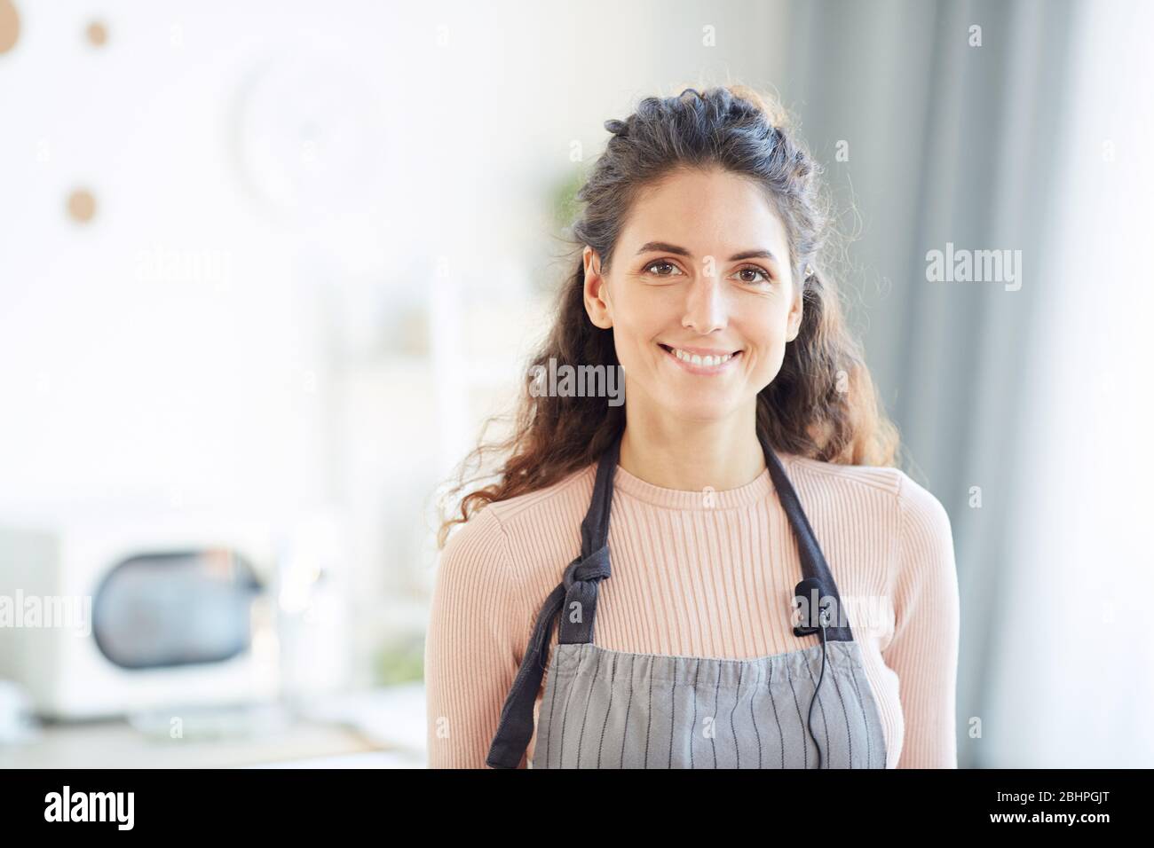 Donna caucasica elegante che indossa grembiule in piedi in cucina guardando la macchina fotografica sorridente, orizzontale petto su colpo Foto Stock