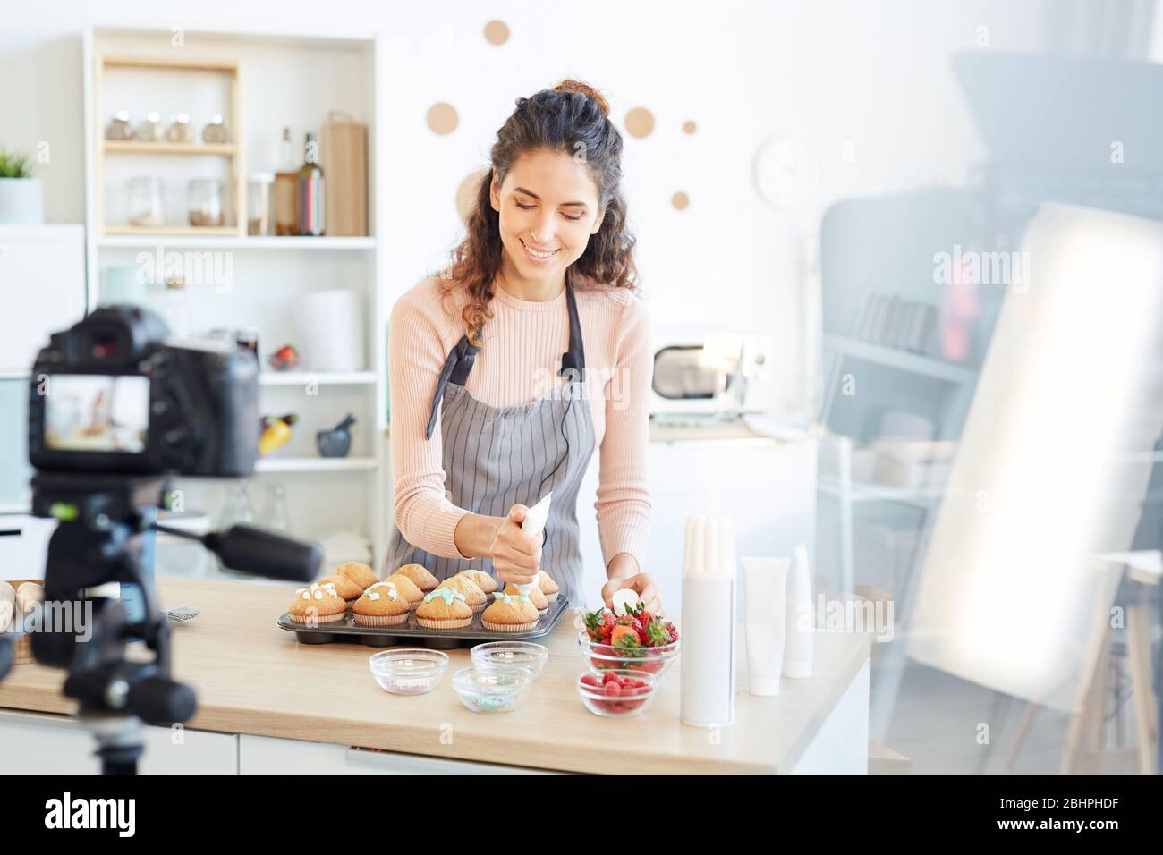 Panettiere professionale in piedi a casa cucina che decorano cupcake freschi con crema dolce, processo di ripresa sulla macchina fotografica Foto Stock
