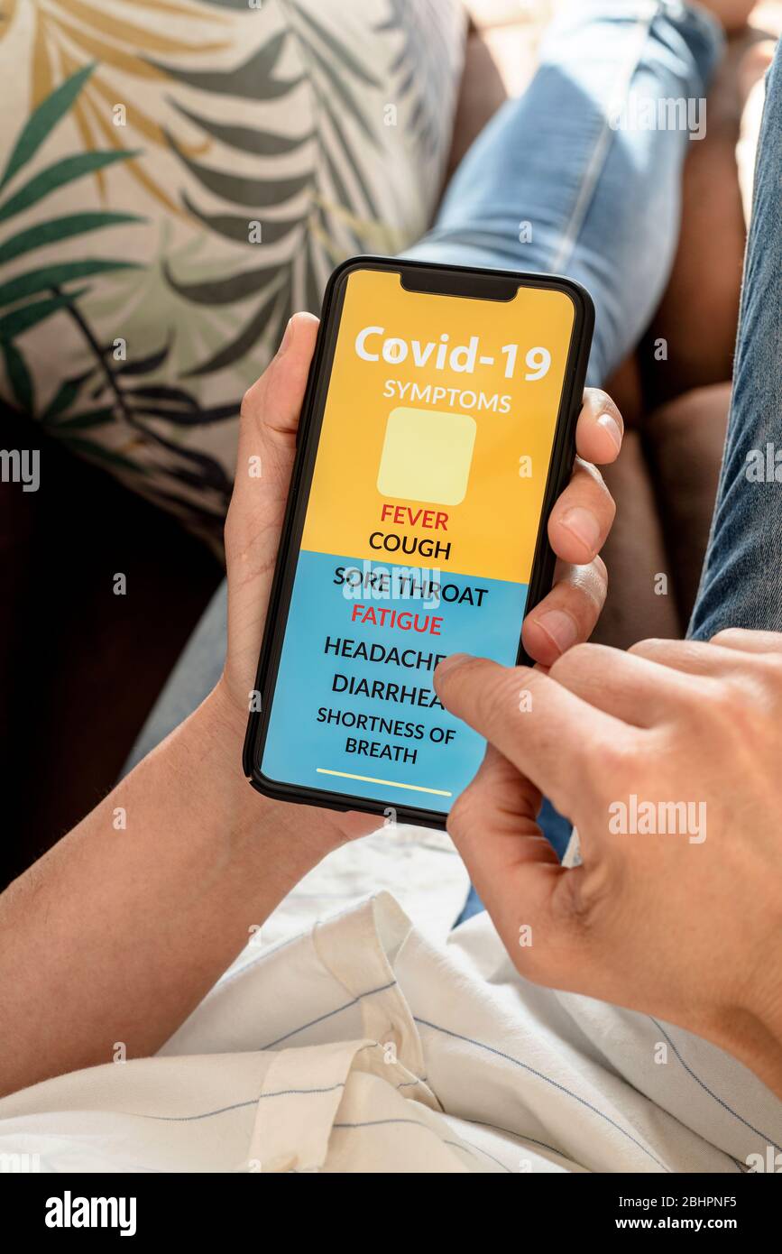 primo piano di un giovane caucasico, comodamente sdraiato su un divano, controllando alcuni dei sintomi del covid-19 in una app simulata nel suo smartphone Foto Stock